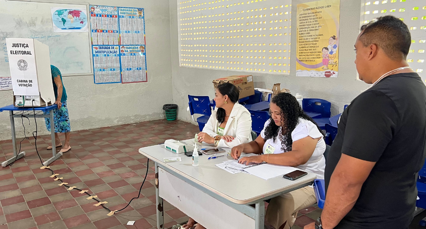 A imagem mostra uma seção eleitoral instalada em uma sala de aula. À esquerda, uma eleitora depo...