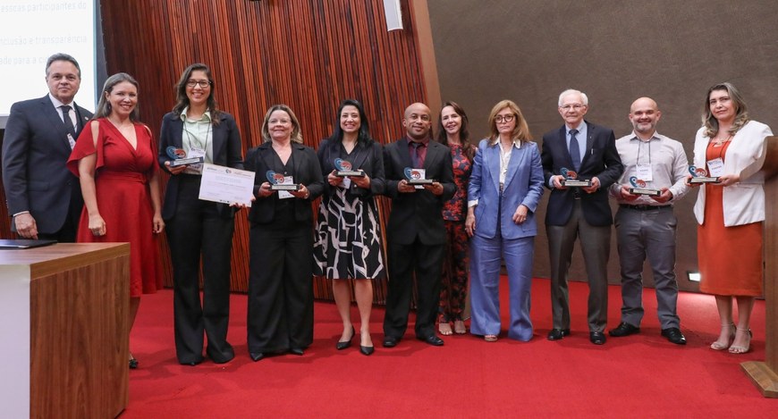 Na foto, um grupo de onze pessoas está posicionado lado a lado no palco, todas voltadas para a c...