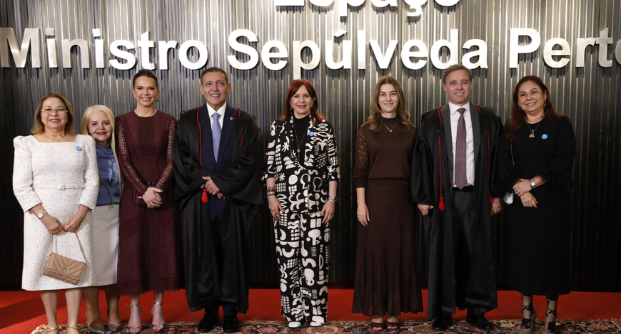 A imagem mostra oito pessoas em pé, posicionadas lado a lado sobre um tapete estampado. Entre el...