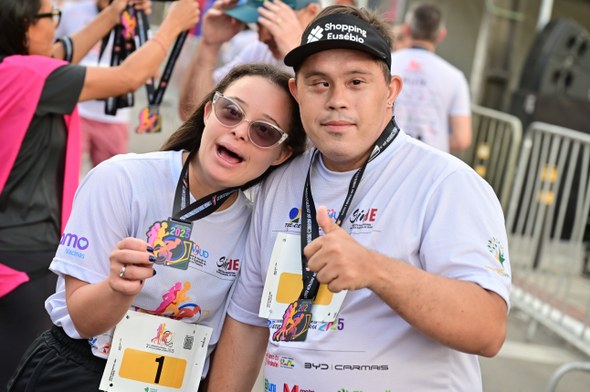 Na sexta imagem, dois participantes posam lado a lado com a medalha do evento. Uma delas é a emb...