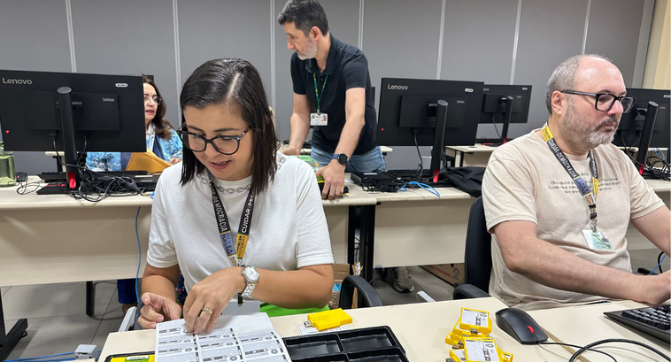 Em uma sala com computadores, servidora de blusa branca e calça verde organiza papéis, mídias e ...