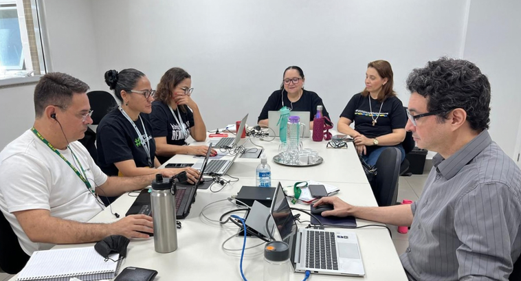 Foto da sala em que foi transmitida a "live". Quatro servidoras e dois servidores estão à mesa. ...