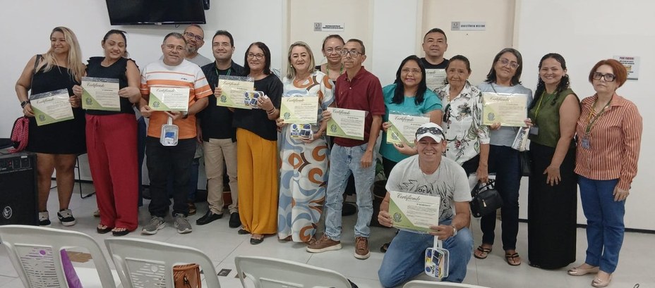 A imagem mostra um grupo de pessoas reunidas em pé e uma ajoelhada em primeiro plano, em uma sal...