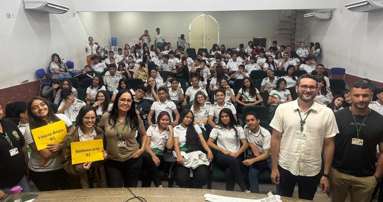 Na imagem, há um auditório com várias fileiras de cadeiras ocupadas por estudantes usando unifor...