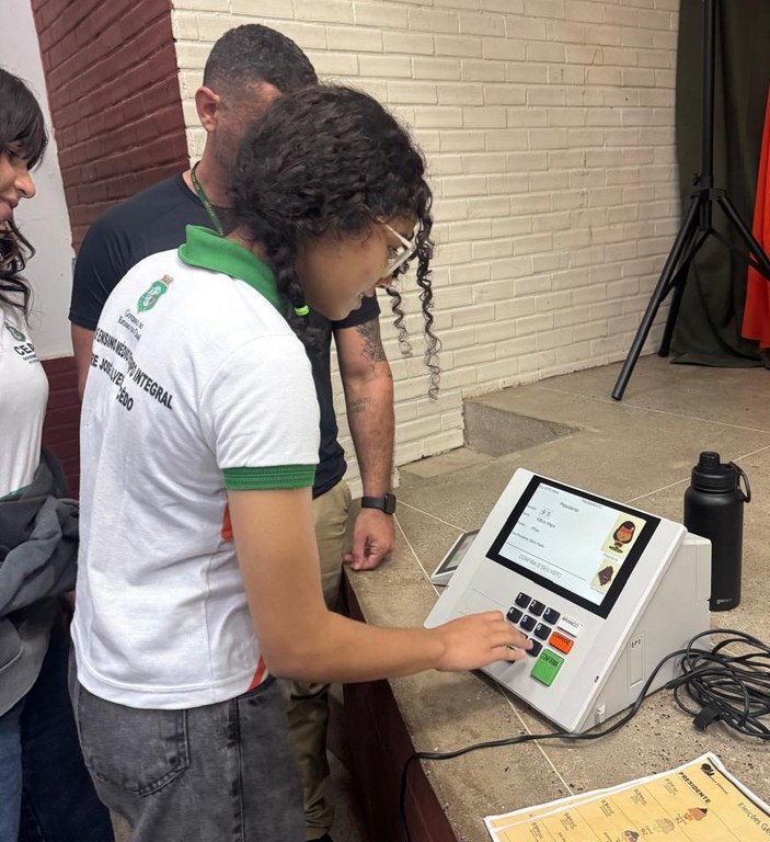 Na imagem, uma estudante com uniforme escolar está em frente a um equipamento eletrônico de vota...