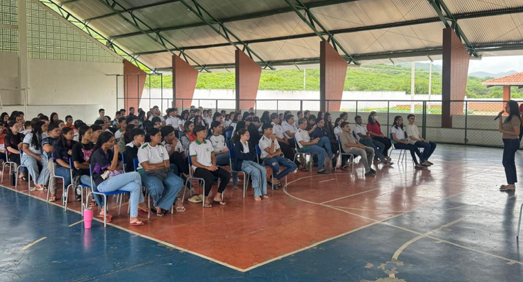 Na imagem, estudantes estão sentados em cadeiras organizadas em filas em uma quadra coberta de e...