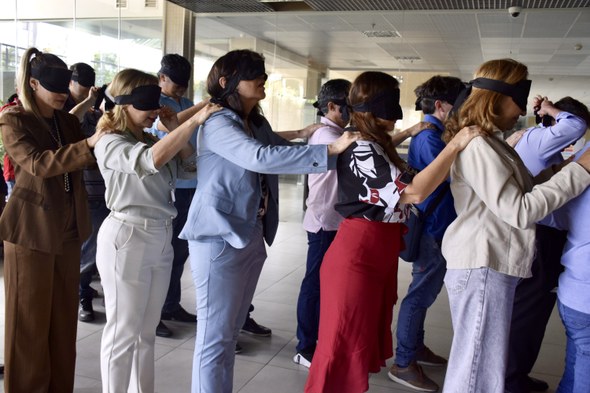 A imagem, também no hall, mostra os participantes enfileirados em duas filas, vendados, com as m...