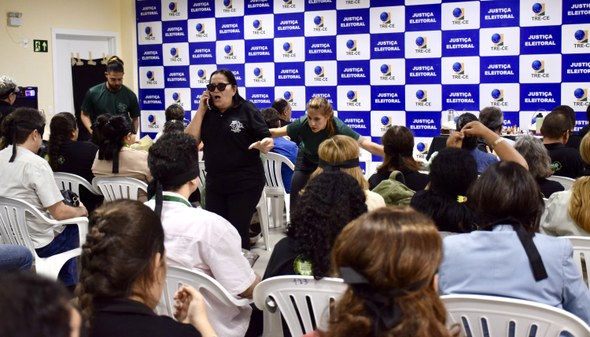 A imagem mostra integrantes de um grupo de teatro atuando entre as fileiras da sala. Ao centro, ...