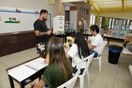 A imagem mostra um ambiente interno de um local de votação. Em primeiro plano, três pessoas estã...