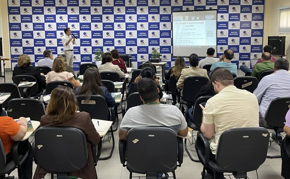 Na imagem, em primeiro plano, os participantes estão sentados em cadeiras, voltados para o pales...