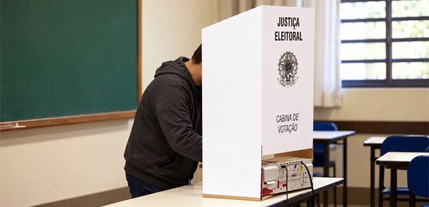 A fotografia mostra uma pessoa em pé, de perfil, votando atrás de uma cabina de votação de papel...