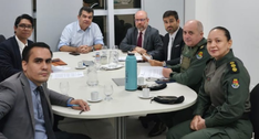 A fotografia registra sete pessoas sentadas ao redor de uma mesa em uma sala de reuniões com par...