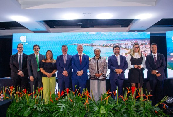 Na foto, nove pessoas posam para a foto em cima de um palco, sendo seis homens e três mulheres. ...