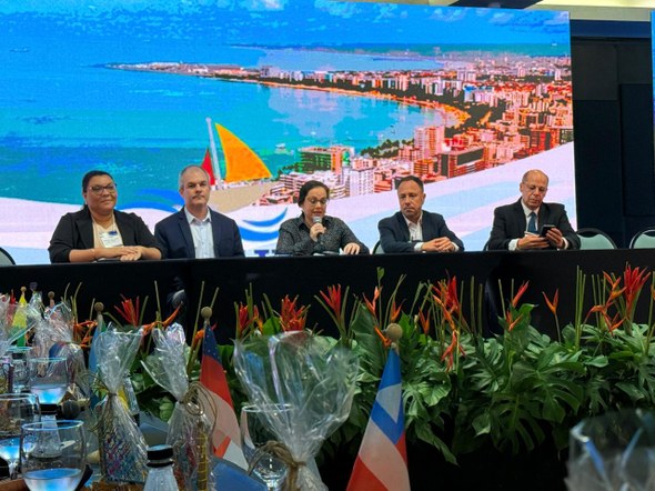 Na foto, cinco pessoas estão sentadas atrás de uma bancada, sendo três homens e duas mulheres. A...