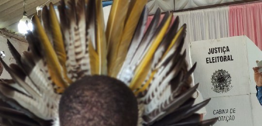 A imagem mostra, em primeiro plano, a parte de trás da cabeça de uma pessoa, que usa um cocar co...