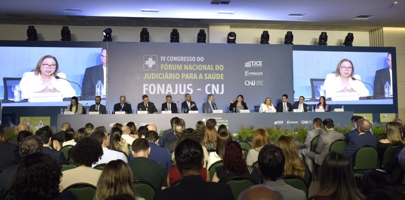 A imagem mostra o auditório do evento. No palco, há uma mesa composta por doze autoridades - oit...
