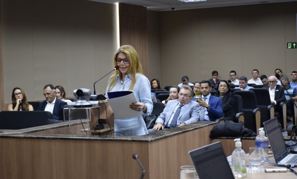 A imagem mostra a juíza Ouvidora da Mulher, Valéria Carneiro, discursando em um púlpito. Ela seg...