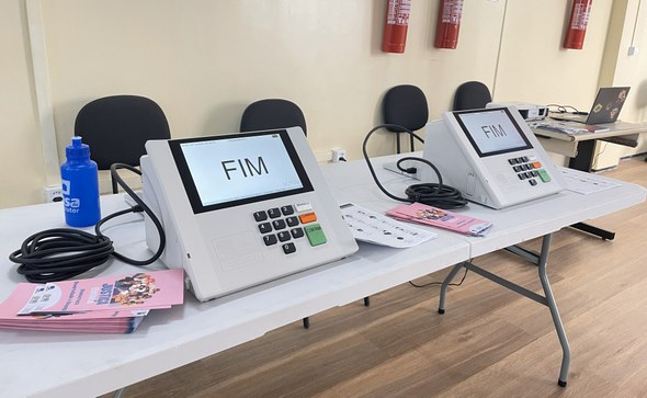 Na imagem, duas urnas eletrônicas estão ligadas sobre uma mesa branca. Nas telas das urnas, apar...