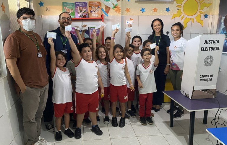 Na imagem, outro grupo de crianças com o mesmo uniforme aparece ao lado de quatro adultos. As cr...