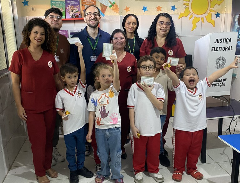 Na imagem, um grupo menor de crianças está à frente e adultos estão atrás. As crianças seguram c...