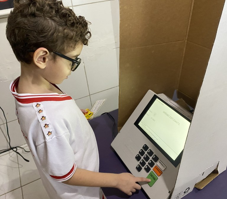 Na imagem, um menino com uniforme escolar está diante de uma urna eletrônica posicionada dentro ...