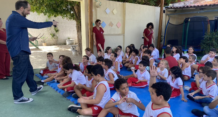 Na imagem, várias crianças com uniforme escolar estão sentadas no chão sobre tapetes em área ext...