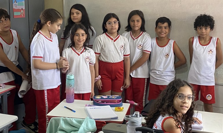 Na imagem, um grupo de crianças com uniforme escolar está dentro de uma sala de aula. Parte das ...