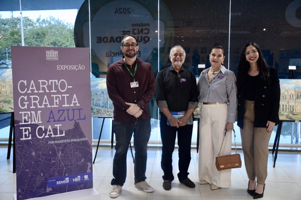 Na imagem há quatro pessoas - duas mulheres e dois homens - em pé, lado a lado, ao lado de uma p...