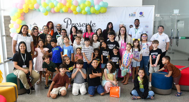 Na imagem, um grupo composto por adultos e crianças está reunido de pé e sentado no chão e em pu...