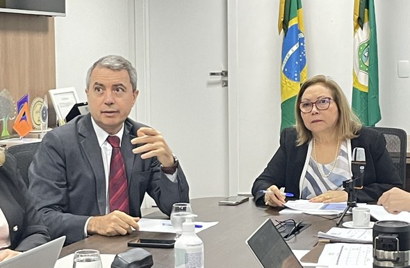 No registro, Maria Iraneide e Edilberto Oliveira estão sentados em torno de uma mesa de madeira,...