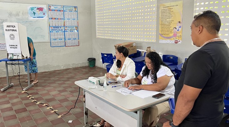 A imagem mostra uma seção eleitoral instalada em uma sala de aula. À esquerda, uma eleitora depo...