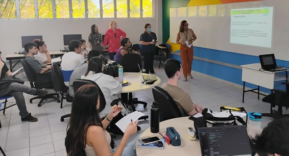Laboratório de Inovação do TRE-CE inicia atividades no campus da Unifor No registro, os estudantes estão sentados, distribuídos em mesas. Em pé, servidoras do TRE-CE fa...