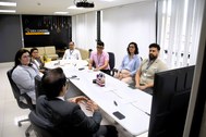 Registro fotográfico feito durante a reunião. Há uma sala com paredes brancas e uma mesa grande ...