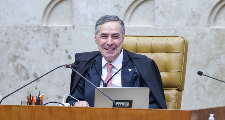 A imagem mostra o ministro Luís Roberto Barroso sorrindo. Ele veste toga preta e está sentado em...
