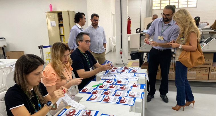 Observadora da Transparência Eleitoral Brasil visita TRE-CE para acompanhar preparação das urnas Um grupo de pessoas trabalha ao redor de uma mesa comprida em um salão com piso claro e paredes ...