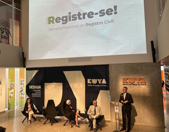 Ouvidor do TRE-CE participa da abertura da 4ª Semana Nacional do Registro Civil, em Fortaleza Na imagem, há um palco com quatro autoridades sentadas em cadeiras e uma em pé, falando ao micro...