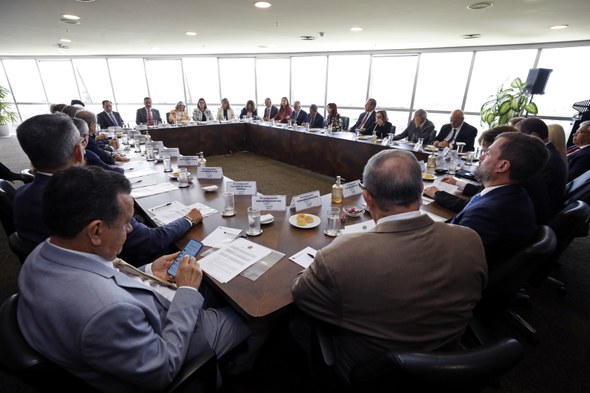 Na imagem, há uma bancada grande de madeira em formato de quadrado fechado. Ao redor dele, os pr...