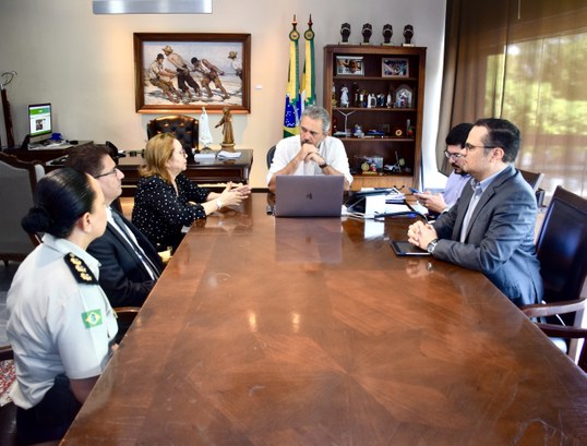 Na imagem, seis pessoas — quatro homens e duas mulheres — estão sentadas em cadeiras em torno de...