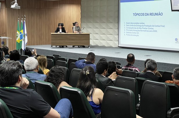 A imagem mostra a visão da plateia em um auditório durante uma palestra. Em primeiro plano, vári...