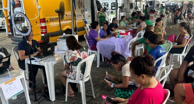 Registro fotográfico feito durante a ação. Nele, há mesas distribuídas em uma praça. Sobre elas,...