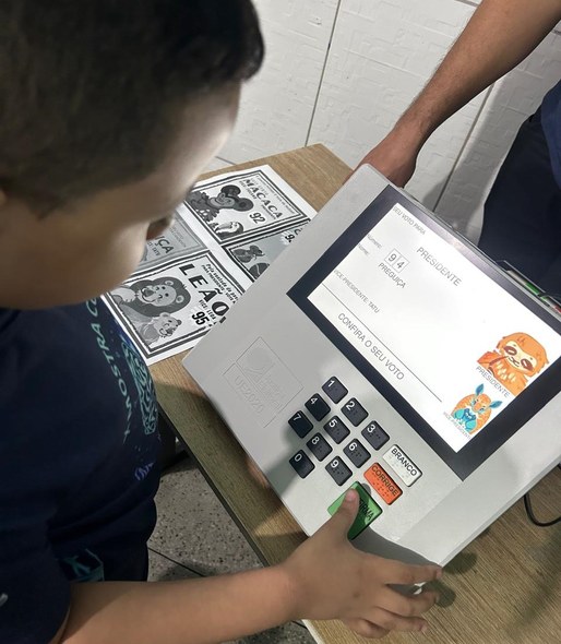 Na imagem, uma criança está em pé diante de uma urna eletrônica posicionada sobre uma mesa de ma...