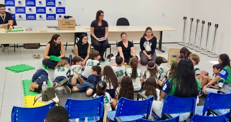 Na foto, um grupo de crianças está sentado no chão de uma sala ampla, organizada com tatames ver...