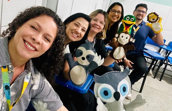 Na imagem, cinco servidores estão sentadas lado a lado em uma fileira de cadeiras azuis, posando...