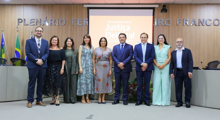 Na imagem, dez pessoas estão posicionadas lado a lado, formando uma fileira à frente da mesa dir...
