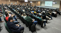 Na imagem, mostra o auditório com participantes sentados em fileiras de cadeiras voltadas para o...