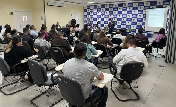 Na imagem, aparece o público em primeiro plano, sentado de costas e voltado para a parte frontal...