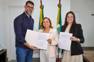 A imagem mostra três pessoas em pé, lado a lado, sorrindo para a câmera em uma sala com paredes ...