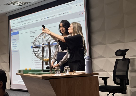 A imagem mostra duas mulheres posicionadas atrás de uma mesa de madeira em no auditório do TRE-C...