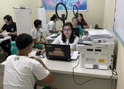A imagem mostra uma sala com mesas organizadas para atendimento. No primeiro plano, um adolescen...
