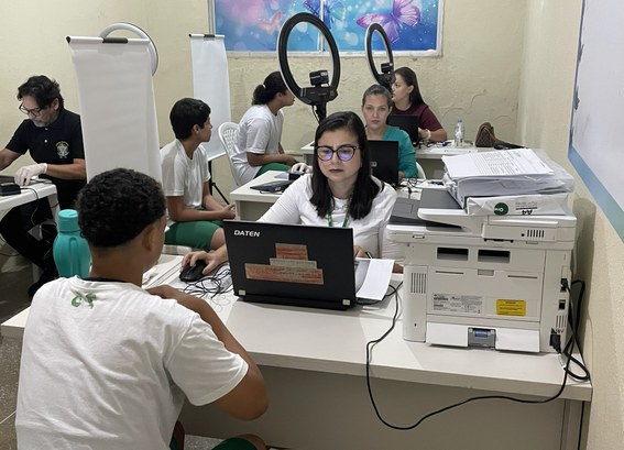 A imagem mostra uma sala com mesas organizadas para atendimento. No primeiro plano, um adolescen...